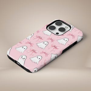 Pode incluir: Capa de telefone rosa com um padrão repetido de fantasmas de desenhos animados brancos e laços rosa. A capa tem uma borda preta e um recorte para a câmera. O design é caprichoso e adequado para o Halloween ou uma estética fofa.