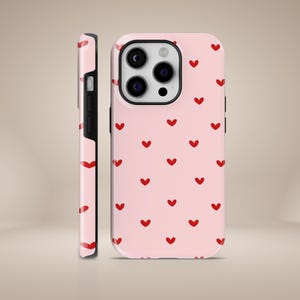 Puede incluir: Una funda de teléfono rosa con un patrón de pequeños corazones rojos. La funda tiene un recorte para el objetivo de la cámara.