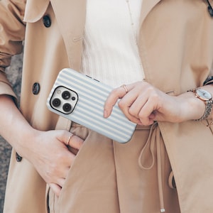 May include: A light blue and white striped phone case. The case is held in front of a beige trench coat. The phone case has a camera and flash cutout on the back.