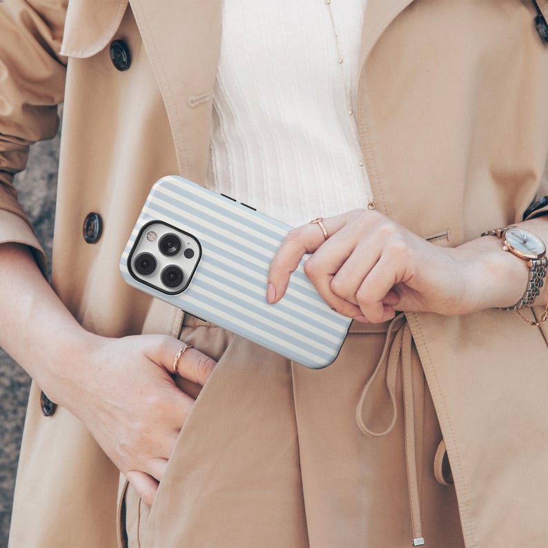 May include: A light blue and white striped phone case. The case is held in front of a beige trench coat. The phone case has a camera and flash cutout on the back.