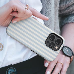 May include: A smartphone case featuring a light blue and white striped design. The case has a black camera module with three lenses. The case is held by a person wearing rings and a watch with a rose gold strap.