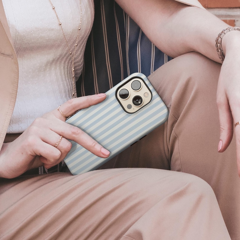 May include: A light blue and white striped phone case. The case is held by a person wearing rings and a bracelet. The phone case has a camera and flash cutout.