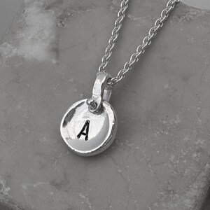 May include: A silver-toned necklace featuring a circular pendant with a slightly irregular, hammered edge. The pendant is stamped with the uppercase letter A in a dark, recessed font. The pendant hangs from a delicate silver-toned chain link.