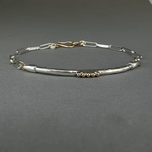 May include: A silver bracelet with a hammered texture and a gold clasp. The bracelet features a series of small gold beads and a chain-link design. The bracelet is displayed on a gray surface.