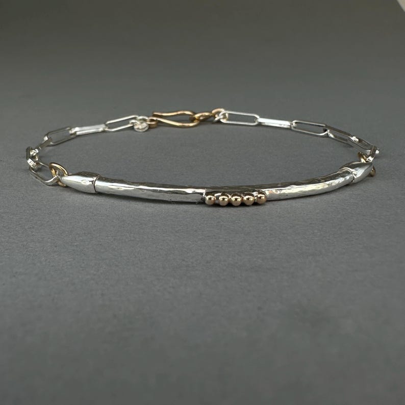May include: A silver bracelet with a hammered texture and a gold clasp. The bracelet features a series of small gold beads and a chain-link design. The bracelet is displayed on a gray surface.
