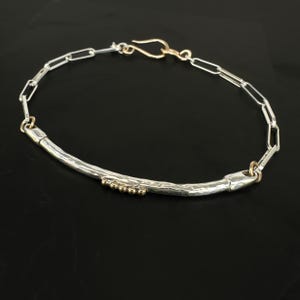 May include: A silver bracelet with a hammered bar design and gold accents. The bracelet features a chain link design with a gold clasp. Small gold beads are set into the silver bar. The jewelry is displayed on a black surface.