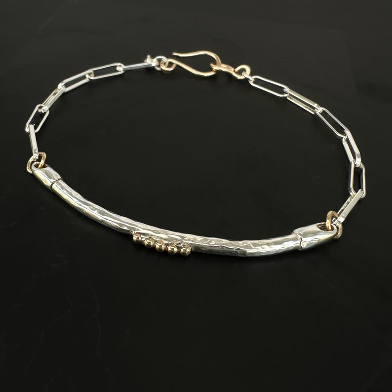 May include: A silver bracelet with a hammered bar design and gold accents. The bracelet features a chain link design with a gold clasp. Small gold beads are set into the silver bar. The jewelry is displayed on a black surface.