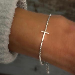 May include: A silver chain bracelet with a delicate cross charm. The bracelet has an adjustable clasp.