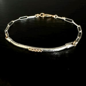 May include: A silver and gold bracelet with a hammered bar and chain link design. The bracelet features a curved silver bar with small gold beads and a gold clasp. The chain links are silver. The jewelry is displayed on a black surface.
