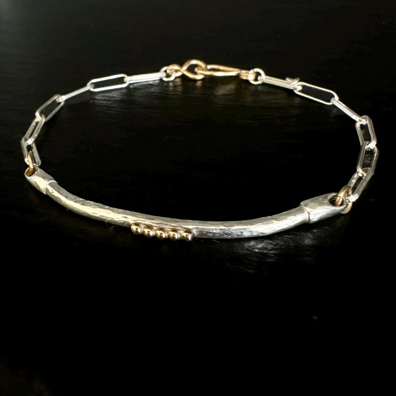 May include: A silver and gold bracelet with a hammered bar and chain link design. The bracelet features a curved silver bar with small gold beads and a gold clasp. The chain links are silver. The jewelry is displayed on a black surface.