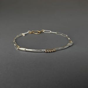 May include: Silver bracelet with a hammered bar and chain link design. The bracelet features gold-colored accents, including a hook clasp and small beads. The jewelry is displayed against a dark gray background.