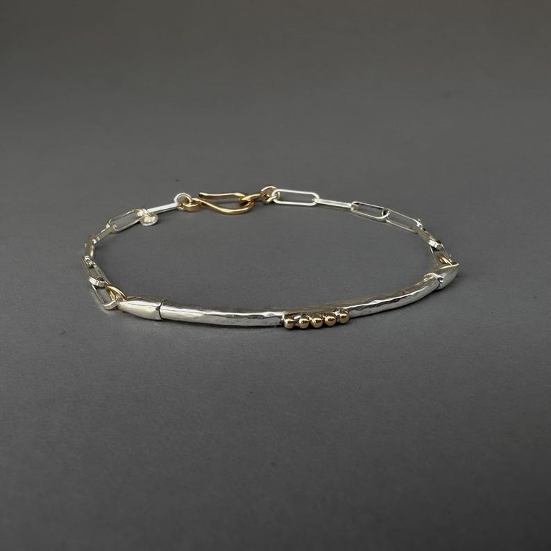 May include: Silver bracelet with a hammered bar and chain link design. The bracelet features gold-colored accents, including a hook clasp and small beads. The jewelry is displayed against a dark gray background.