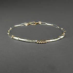 May include: A silver bracelet with a hammered bar and gold accents. The bracelet features a chain link design with a gold clasp. The bar has three small gold beads. The jewelry is displayed on a gray background.