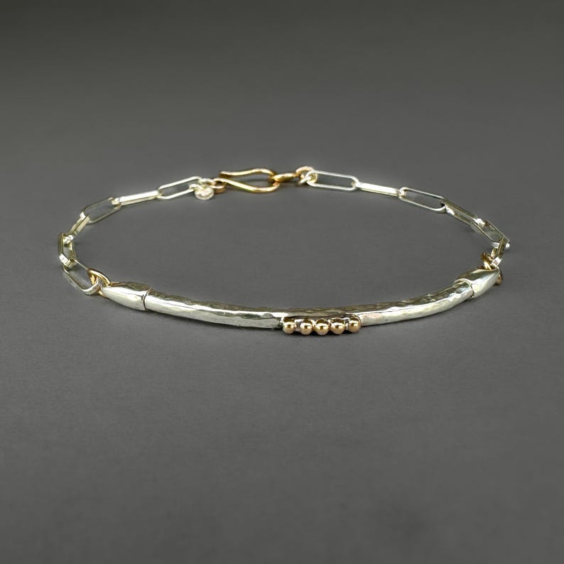May include: A silver bracelet with a hammered bar and gold accents. The bracelet features a chain link design with a gold clasp. The bar has three small gold beads. The jewelry is displayed on a gray background.