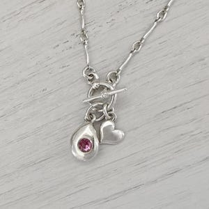 May include: A silver necklace featuring a toggle clasp closure with two dangling charms. One charm is a smooth, rounded teardrop shape set with a single round pink gemstone, and the other charm is a simple, polished silver heart shape.