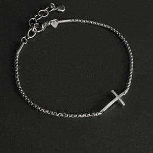 May include: A silver-colored bracelet with a cross pendant. The bracelet features a delicate chain design and an adjustable clasp. The cross is positioned on the side of the bracelet. The background is a dark gray.