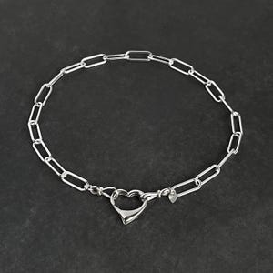 May include: A silver chain necklace with a heart-shaped clasp. The chain is made of small, oval-shaped links.