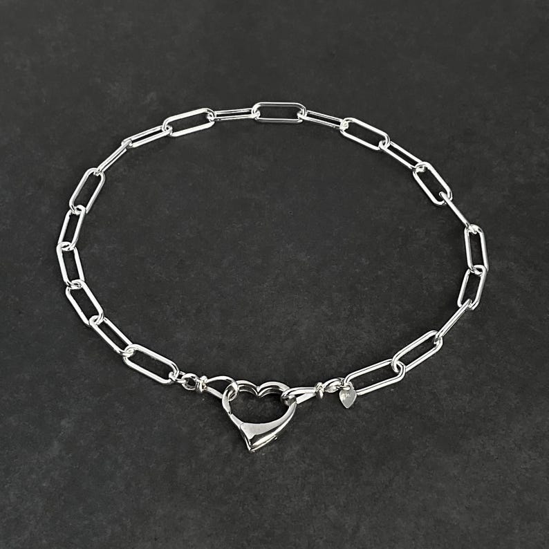 May include: A silver chain necklace with a heart-shaped clasp. The chain is made of small, oval-shaped links.