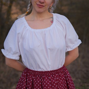 May include: A white peasant blouse with a gathered neckline and short sleeves, paired with a burgundy skirt with a white floral pattern.