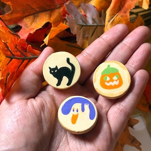Puede incluir: Tres pequeños discos de madera redondos y de color beige con diseños de Halloween pintados a mano. Un disco muestra un gato negro, otro una calabaza naranja con tallo verde y el tercero un fantasma blanco con detalles morados.