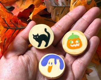 May include: Three small, round, beige wooden discs featuring hand-painted Halloween designs. One disc displays a black cat, another shows an orange jack-o'-lantern with a green stem, and the third features a white ghost with purple accents.
