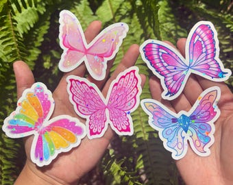 Può includere: Cinque adesivi fustellati a forma di ali di farfalla con finiture olografiche. Un adesivo è color arcobaleno, uno è rosa chiaro, uno è rosa acceso con motivi dettagliati, uno è magenta e blu, e uno è blu e viola.