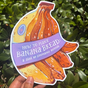 May include: A die-cut sticker shaped like a bunch of bananas with a purple banner across the center. The text reads: HOW TO MAKE BANANA BREAD: a zine BY STEPHANIE HUTTON. The illustration features yellow and orange bananas with dark brown spots.