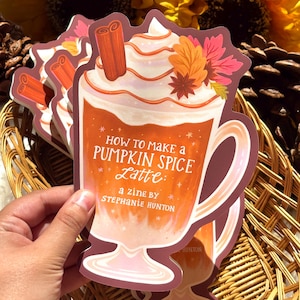 May include: A die-cut sticker shaped like a mug of pumpkin spice latte with whipped cream, cinnamon sticks, and autumn leaves. The text reads: How to make a PUMPKIN SPICE Latte: a zine by STEPHANIE HUNTON. The design features orange and cream colors.
