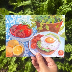 Puede incluir: Una pintura colorida en una tarjeta rectangular que muestra un tazón de tomates rojos, un plato con un huevo frito sobre tostada de aguacate y rodajas de naranja. La obra presenta una escena de desayuno con una planta en maceta.