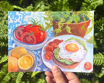 May include: A colorful painting on a rectangular card featuring a bowl of bright red tomatoes, a plate with a fried egg on avocado toast, and sliced oranges. The artwork displays a vibrant breakfast scene with a potted plant in the background.