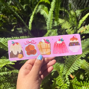 Puede incluir: Un marcap&aacute;ginas rosa con ilustraciones de postres. Los postres incluyen un pastel, galletas, un gofre, una gelatina y una magdalena. El marcap&aacute;ginas se sostiene frente a un fondo de follaje verde.