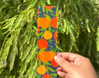 May include: A rectangular bookmark featuring a dark blue background with a repeating pattern of bright orange citrus fruits and green leaves. The design displays multiple oranges in various shades of orange and yellow with green foliage.