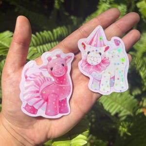 May include: Two stickers with playful animal illustrations. One depicts a pink giraffe with a striped skirt and polka dots. The other shows a white llama wearing a pink party hat and frilly collar. Both stickers have glitter details.