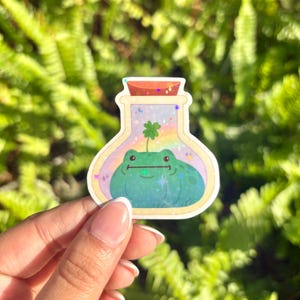 Könnte beinhalten: Ein skurriler Aufkleber mit einem Cartoon-Frosch in einer Zaubertrankflasche. Der Frosch ist grün und lächelt, und ein vierblättriges Kleeblatt wächst aus seinem Kopf. Die Flasche hat einen Regenbogenhintergrund und Glitzerakzente.
