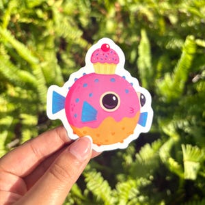 Puede incluir: Una colorida pegatina con un pez globo de dibujos animados con una magdalena en la cabeza. El pez globo es rosa con aletas azules y detalles naranjas. La magdalena es amarilla con chispas rosas y una cereza roja. La pegatina está sujeta por una mano.