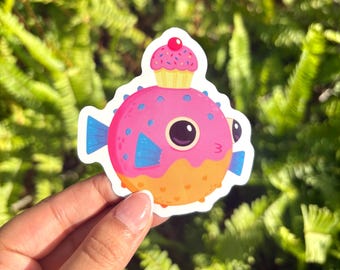May include: A colorful sticker featuring a cartoon pufferfish with a cupcake on its head. The pufferfish is pink with blue fins and orange accents. The cupcake is yellow with pink sprinkles and a red cherry on top. The sticker is held by a hand.