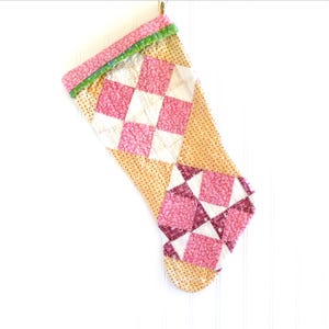 May include: A handmade Christmas stocking with a patchwork design. The stocking features a combination of pink floral and cream-colored fabrics, arranged in a diamond pattern. The top edge is trimmed with green and pink fabric. A gold loop is attached for hanging.