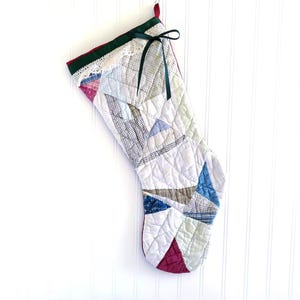 May include: A quilted Christmas stocking with a patchwork design. The stocking features a variety of fabrics in shades of white, blue, green, and burgundy. A green ribbon is tied at the top, and the top edge is trimmed with lace.