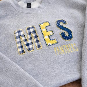 Pode incluir: Um moletom cinza com as letras "MES" aplicadas em tecido azul e amarelo. A palavra "PANTHERS" é bordada em amarelo abaixo das letras. As letras têm uma borda costurada.