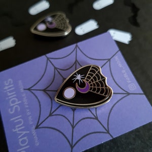May include: Black and white enamel pin shaped like a planchette with a spider, crescent moon, and star. The pin is on a purple background with a spiderweb pattern. The text "Playful Spirits" is printed on the card.