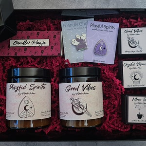 Może przedstawiać: Czarne pudełko prezentowe wypełnione czerwoną strzępioną papierem i różnymi przedmiotami, w tym dwie świece z etykietą "Playful Spirits" i "Good Vibes", małe pudełko z etykietą "Crystal Visions", małe pudełko z etykietą "Moon Tea" i dwie emaliowane szpilki z etykietą "Friendly Ghost" i "Playful Spirits".