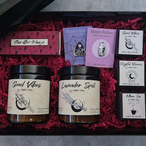 Op de afbeelding: Een zwarte cadeaudoos gevuld met rood geribbeld papier en verschillende heksenthema-artikelen, waaronder kaarsen, emaillepins en een klein doosje met het label "Moon Tea". De kaarsen zijn gelabeld "Good Vibes", "Lavender Spell" en "Crystal Visions". De emaillepins zijn gelabeld "Playful Spirit" en "Moon Potion".