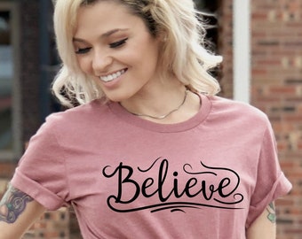 May include: A dusty rose colored t-shirt with the word "Believe" written in black cursive script.