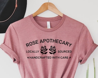 May include: A heather mauve t-shirt with the text "ROSE APOTHECARY" printed in black. The text is surrounded by three black roses. The text below the roses reads "LOCALLY SOURCED HANDCRAFTED WITH CARE".