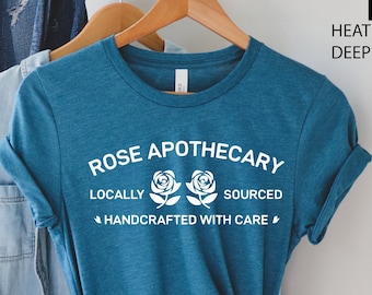 May include: A teal blue t-shirt with the text "Rose Apothecary" in white. The text is surrounded by two white roses and the words "Locally Sourced" and "Handcrafted with Care".