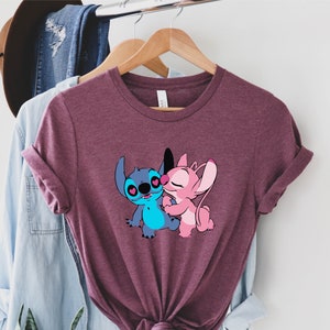 May include: A maroon t-shirt with a graphic of Stitch and Angel from Lilo & Stitch. Stitch is blue and Angel is pink. They are hugging and have hearts for eyes.