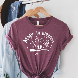 May include: A maroon t-shirt with the text "Magic in progress" in white. The shirt features a graphic of a broom, a pair of glasses, a wand, a baby footprint, and a witch's hat.