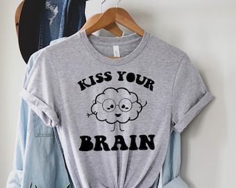 May include: A light gray t-shirt with the phrase "KISS YOUR BRAIN" in black, along with a cartoon brain graphic wearing glasses. The shirt is hanging on a wooden hanger, with a denim shirt and a hat in the background.