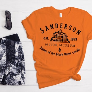 Puede incluir: Camiseta naranja con un dise&ntilde;o gr&aacute;fico negro que presenta una casa, el texto "Sanderson est. 1693 Witch Museum Home of the black flame candle" y un s&iacute;mbolo de llama negra.