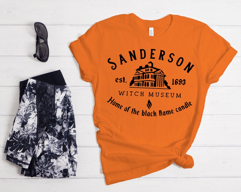 Puede incluir: Camiseta naranja con un dise&ntilde;o gr&aacute;fico negro que presenta una casa, el texto "Sanderson est. 1693 Witch Museum Home of the black flame candle" y un s&iacute;mbolo de llama negra.